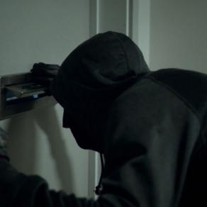 A man wearing dark clothes looks through a door's mail slot. | Still from "Follow the Money" Season 2, Episode 5